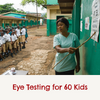 A women holding a stick against a wall with eye testing paper surrounded by kids with the caption saying ' eye testing for 60 kids'